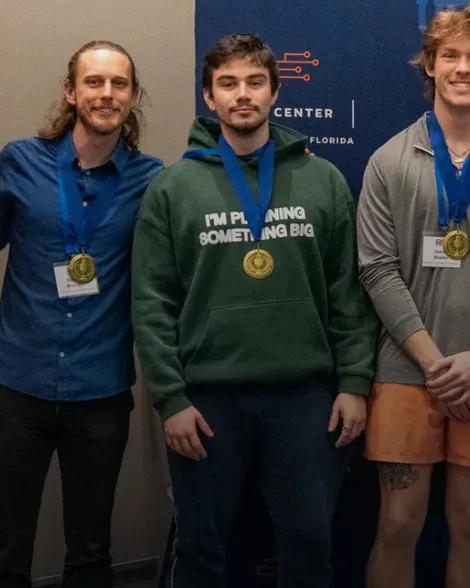 Raul in a green hoodie wearing a GatorHack first-place medal, between two teammates.
